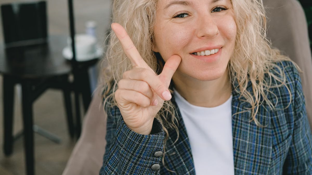 Crop happy adult female entrepreneur with blond hair in formal clothes smiling and showing index finger up sign while looking at camera sitting in armchair in cafe
